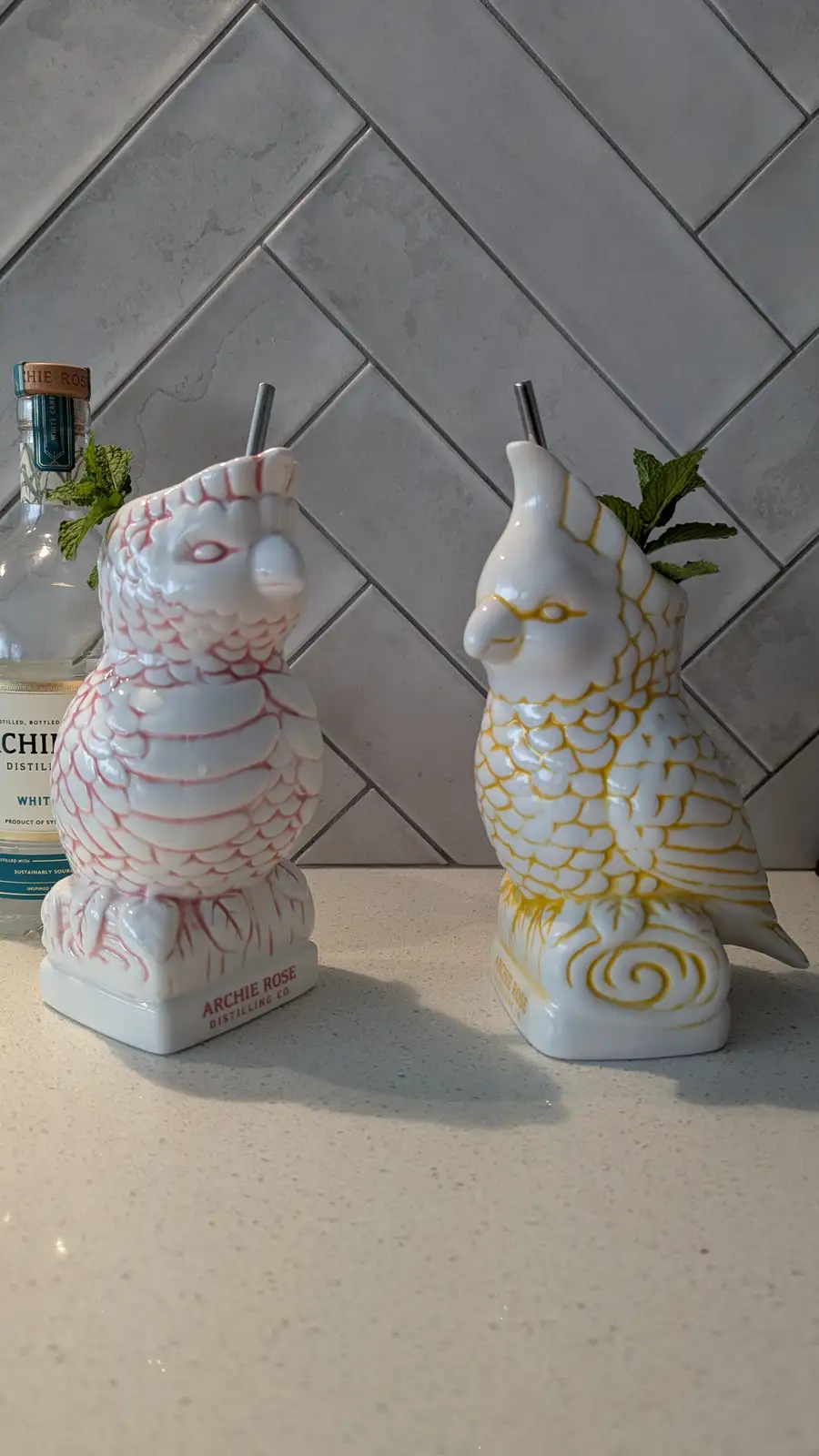 Two ceramic bird-shaped cocktail glasses, one pink and one yellow, sit on a countertop with mint sprigs and straws. A bottle…