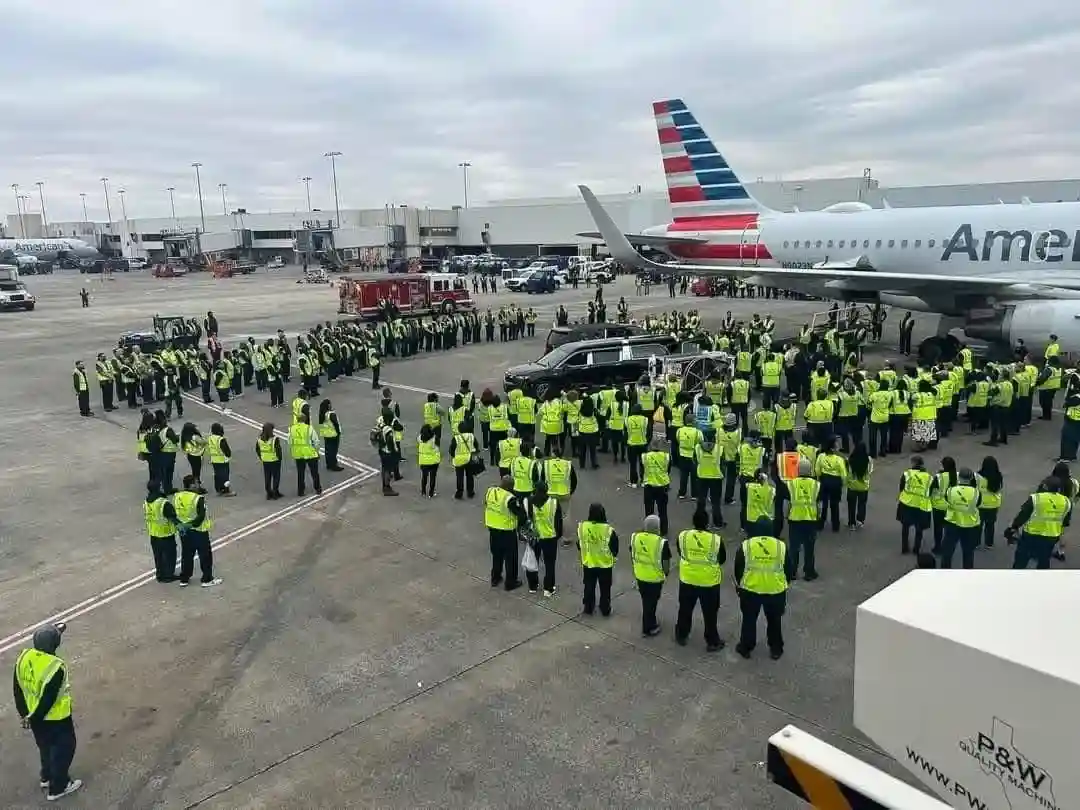 A large group of airport ground crew in bright yellow safety vests stand in formation on an airport tarmac, surrounding a…