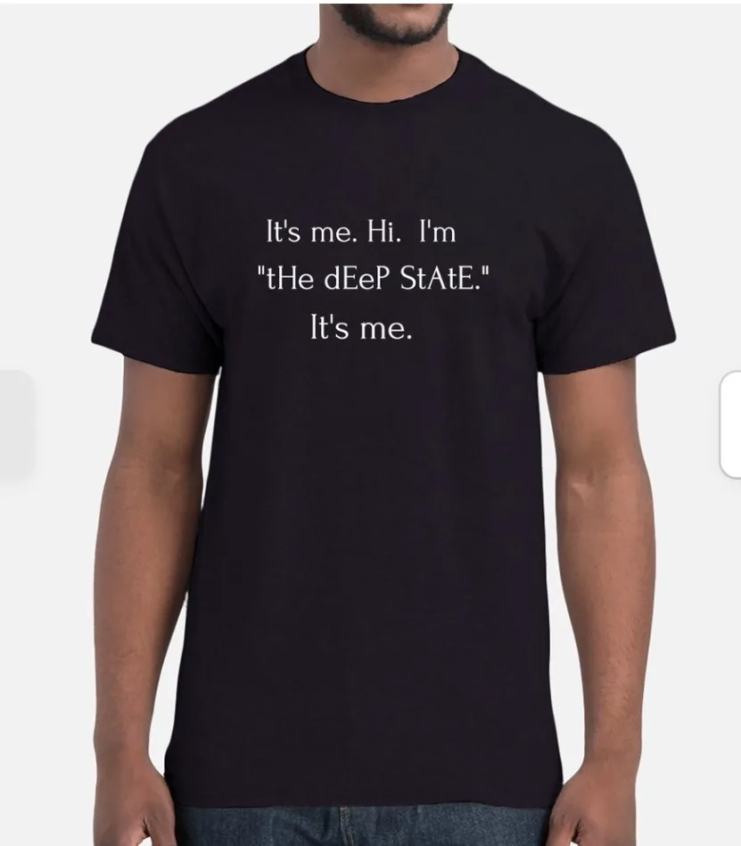 A person wearing a black t-shirt with white text that reads 'It's me. Hi. I'm "tHe dEeP StAtE." It's me.' The text is styled…