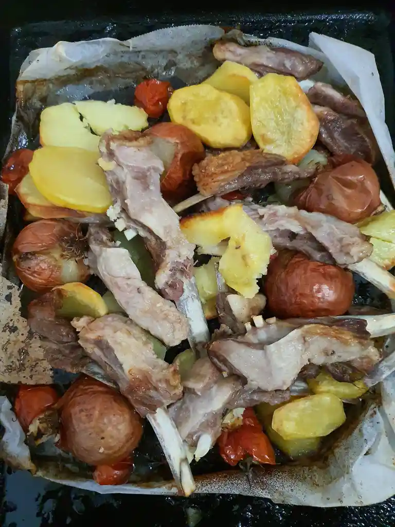 A close-up photo of a baking tray lined with parchment paper, filled with roasted lamb chops, golden-brown potato chunks…