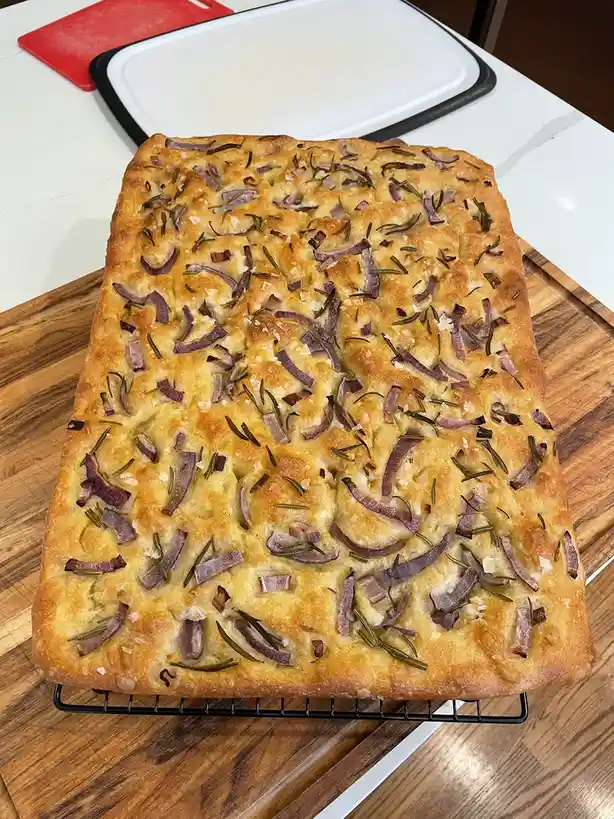 A freshly baked rectangular focaccia bread sits on a black wire cooling rack atop a wooden cutting board. The golden-brown…