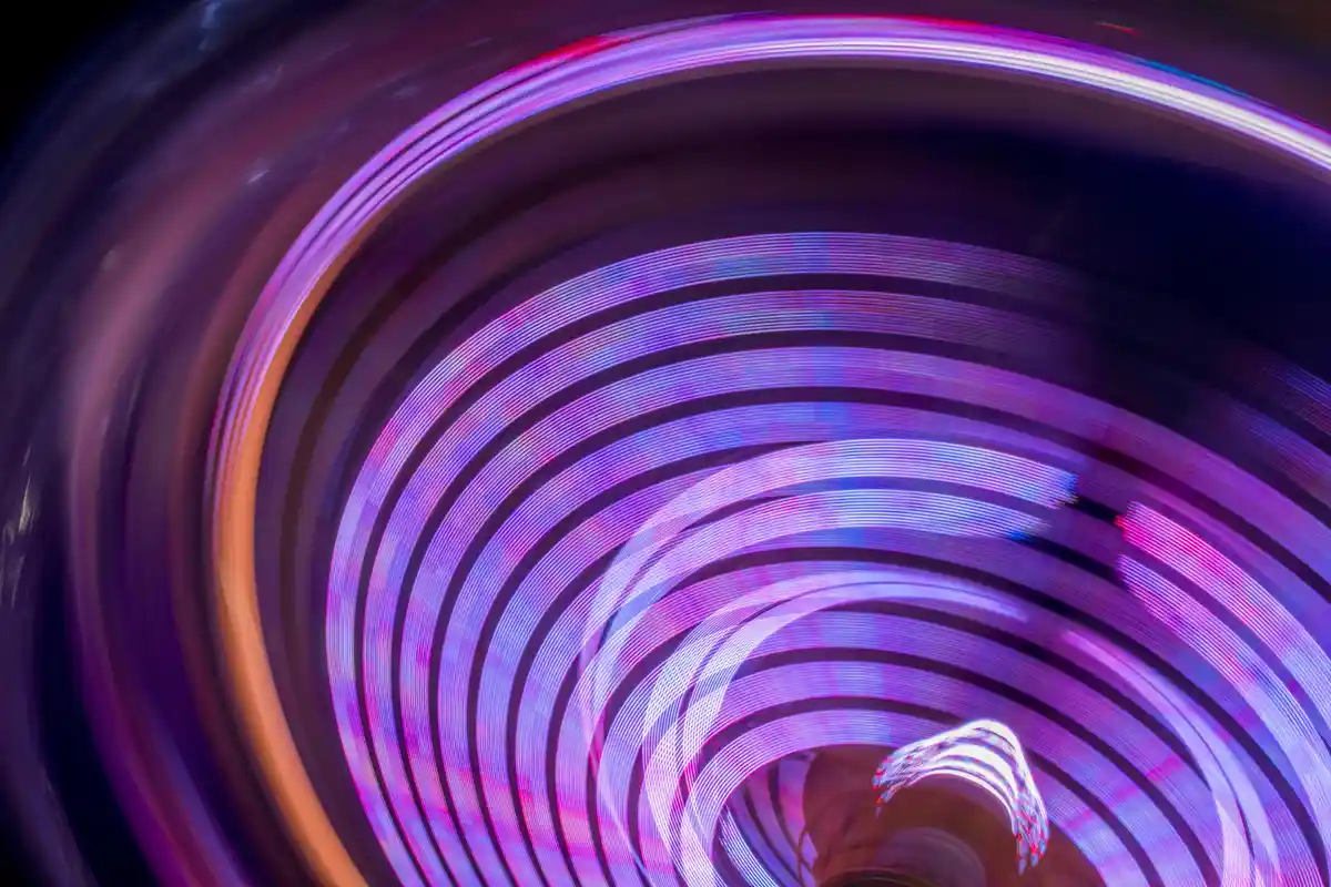 A long-exposure photograph shows swirling, concentric light trails in vibrant purples, pinks, and blues against a dark…