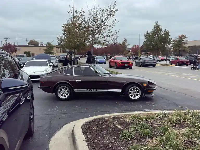 A maroon Datsun 240Z with white racing stripes is parked in a crowded lot filled with other classic and modern cars…