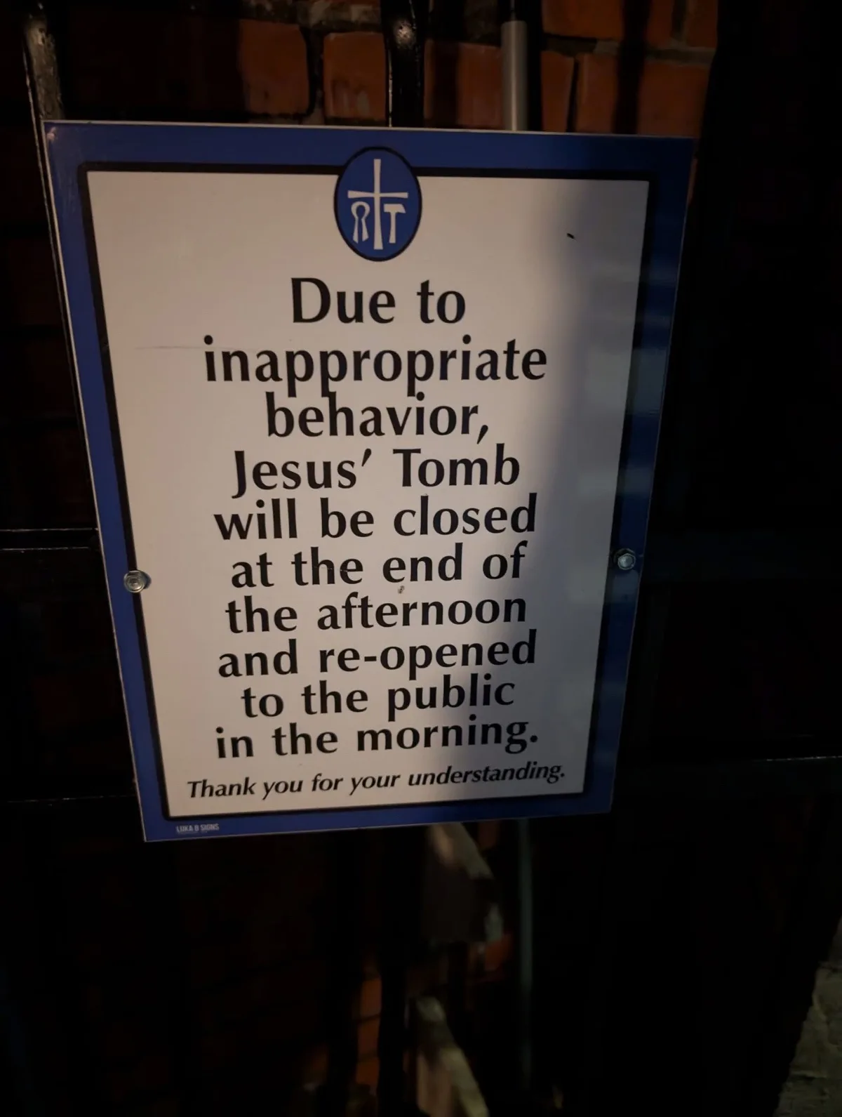 A white sign with a blue border and a religious symbol at the top is mounted on a dark metal gate against a brick wall. The…