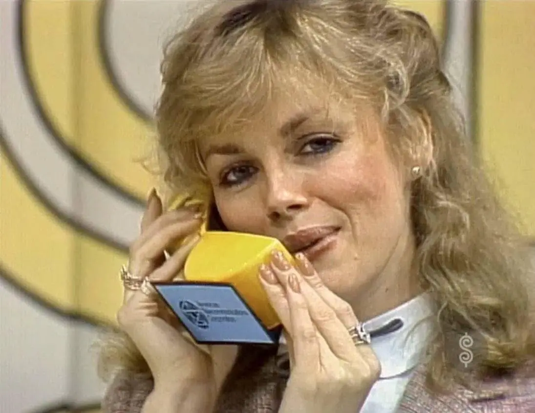 a woman with blonde hair holds a yellow, retro-style telephone receiver to her ear while smiling slightly, her hands…