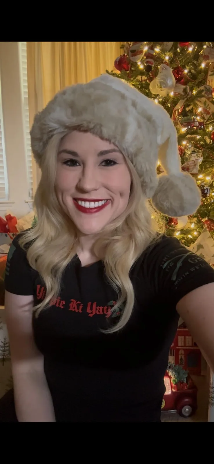 A smiling blonde woman wearing a white furry Santa hat and a black t-shirt with red text that reads 'Yippe Ki Yay' poses in…