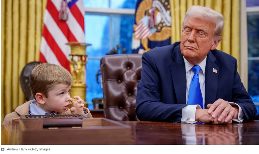 a young boy sits at a desk in the oval office, looking up at former president donald trump who is seated across from him…