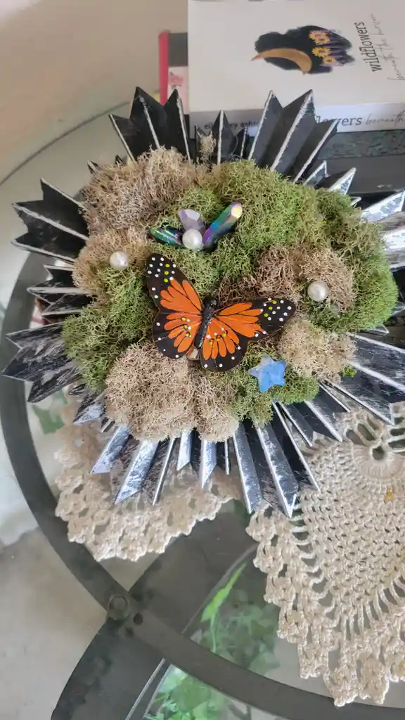 A decorative craft item resembling a sunflower or wreath, made of green and beige moss, black and silver spiked rays, and…