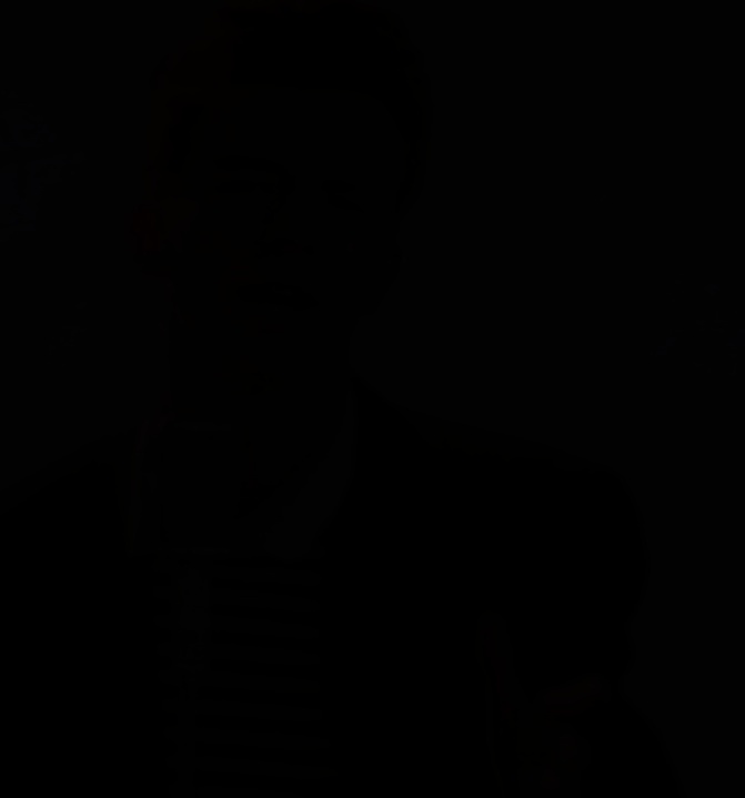 A very dark, almost completely black image with faint, barely visible outlines of a person's face and shoulders. The…