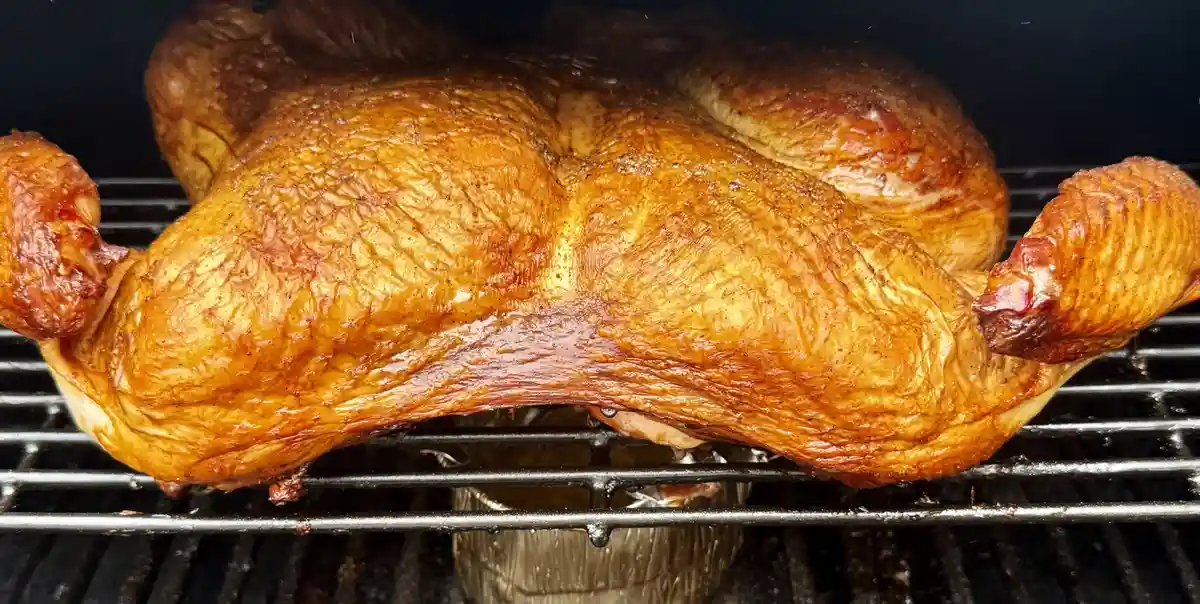 A whole roasted chicken with golden-brown, crispy skin sits on metal grill grates inside a smoker, with visible smoke and a…
