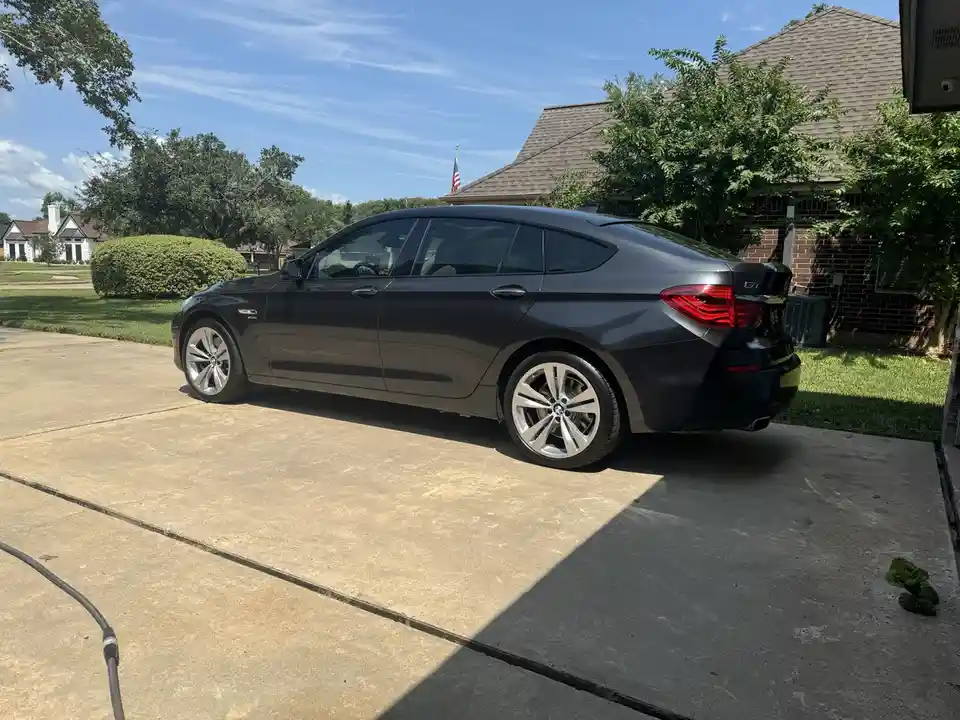 A dark gray BMW GT sedan is parked on a concrete driveway in front of a brick house with an American flag on the roof. The…