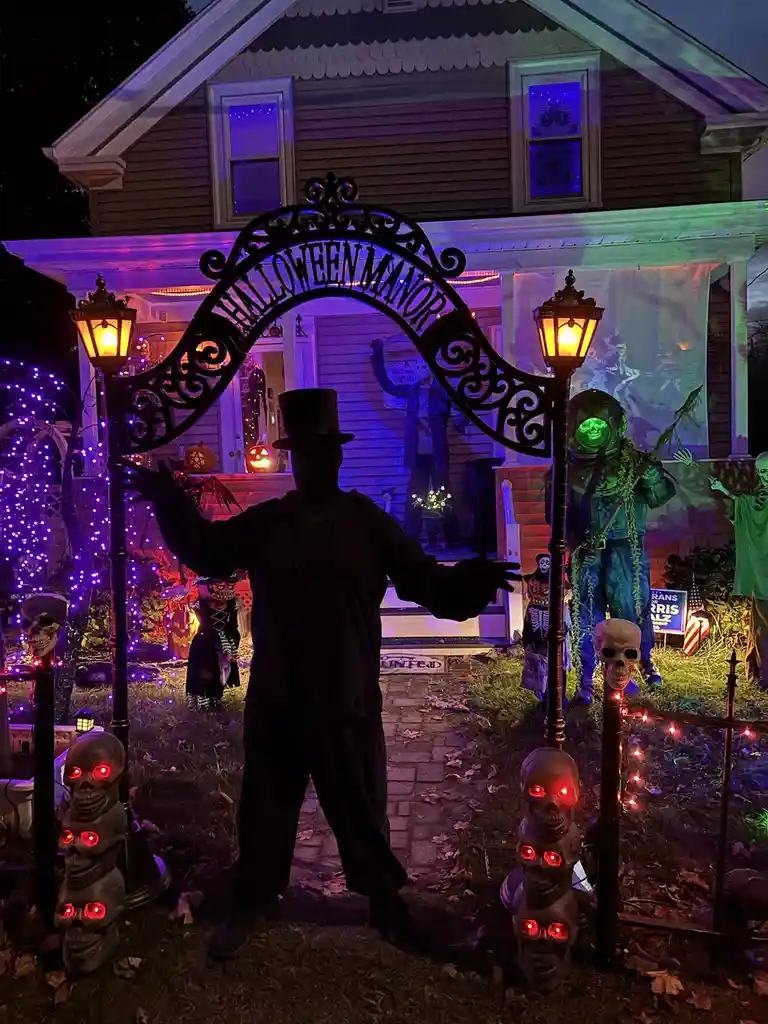 A person in a black top hat and coat stands in silhouette at the entrance of a heavily decorated Halloween house, arms…