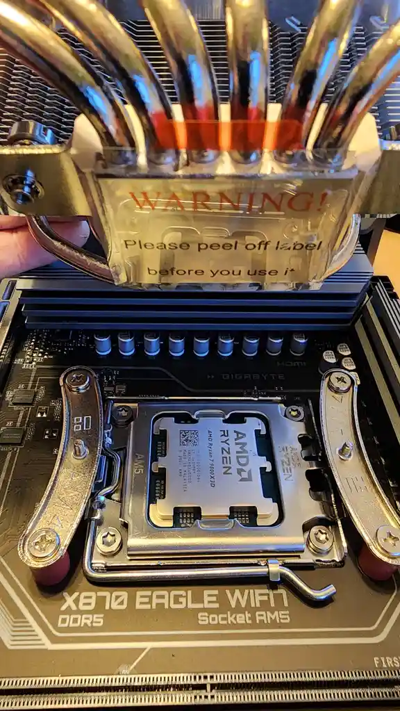 A close-up photo shows a hand holding a CPU cooler over an AMD Ryzen 7 9800X3D processor installed in a Gigabyte X870 Eagle…