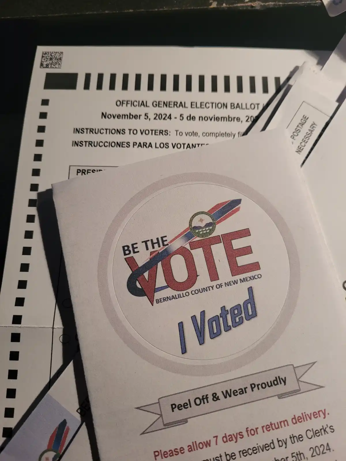 A close-up photo shows a white 'I Voted' sticker from Bernalillo County, New Mexico, featuring the text 'Be the Vote' and 'I…