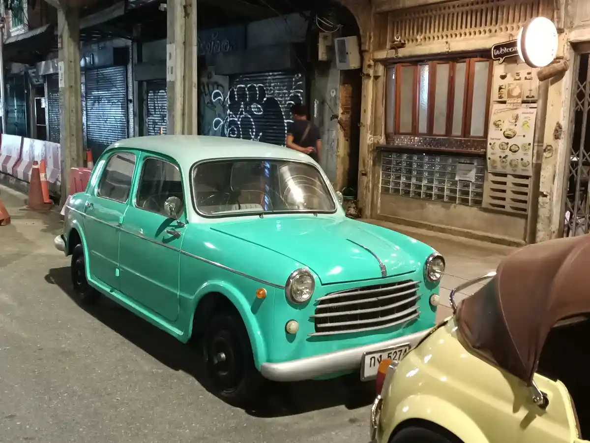 A vintage turquoise car is parked on a street at night, with a yellow car partially visible in the foreground. The…