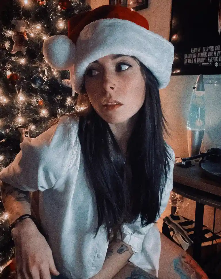 A person with long dark hair and visible tattoos wears a red and white Santa hat while sitting in front of a lit Christmas…