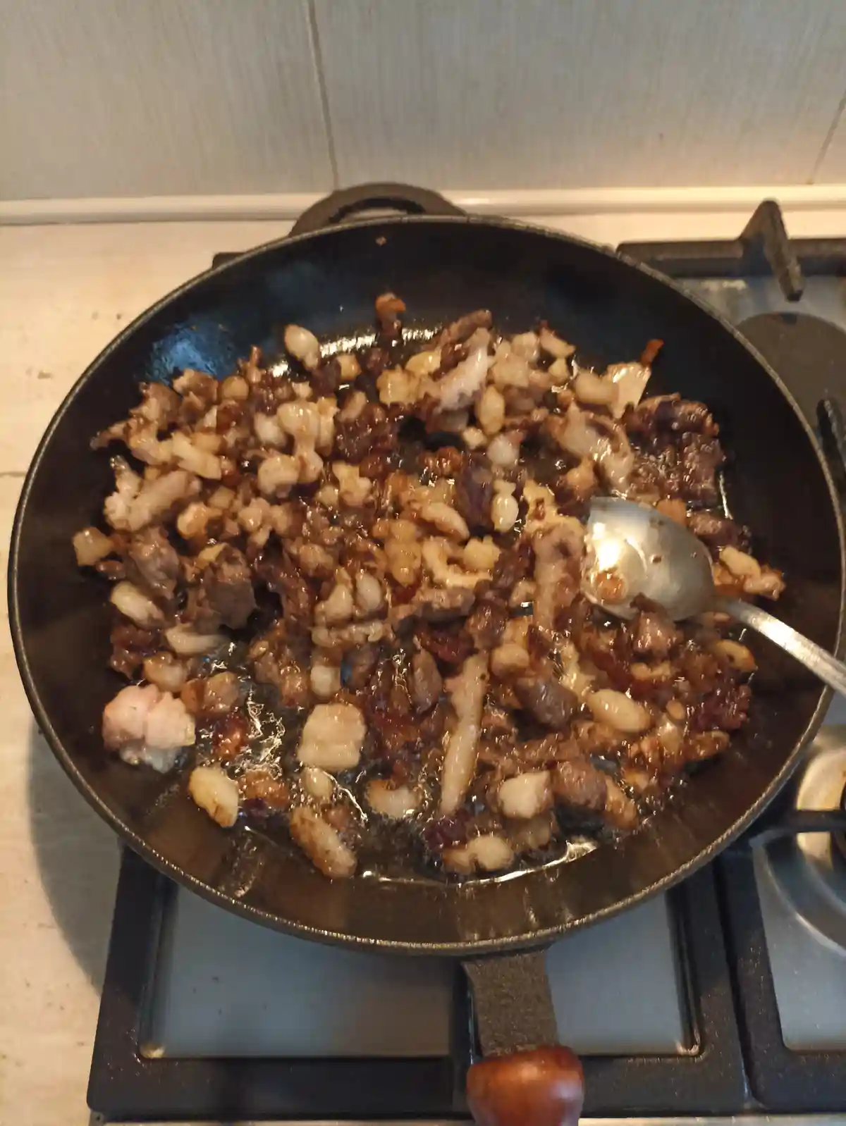 A close-up photo shows a black cast-iron skillet on a gas stove, filled with sizzling, crispy bacon pieces and rendered fat…
