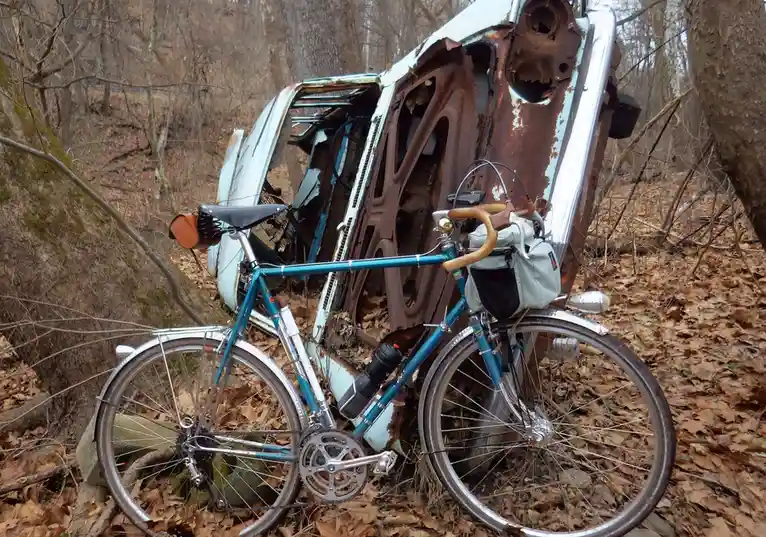 A teal bicycle with leather handlebars and a white bag is leaning against a rusted, overturned light-blue car in a…