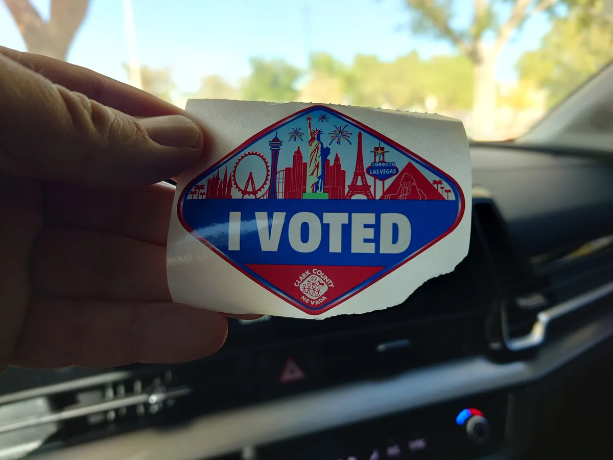 A hand holds up a torn 'I VOTED' sticker from Clark County, Nevada, featuring a collage of global landmarks like the Statue…