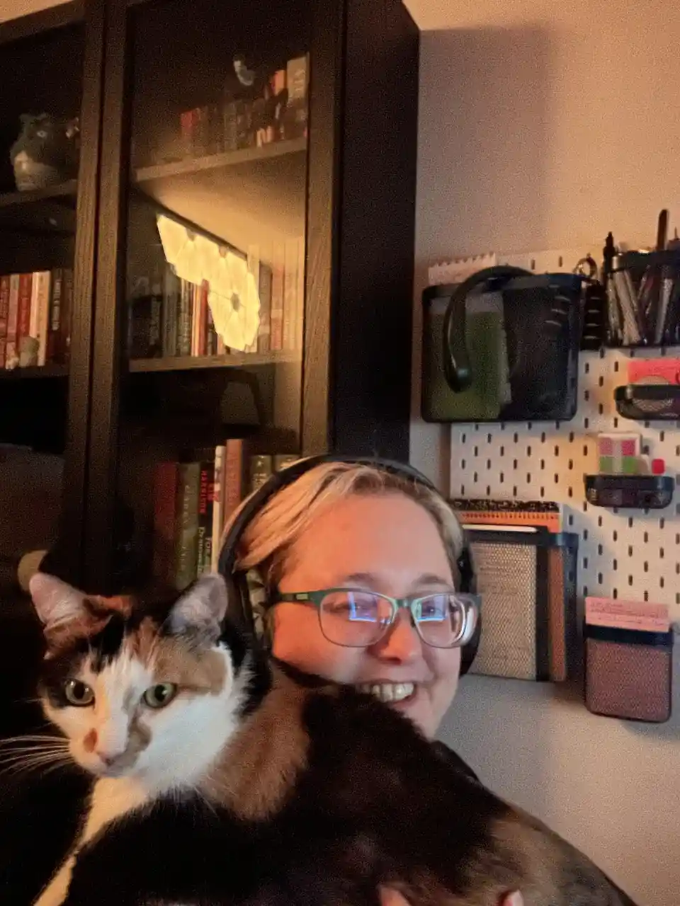 A person with short blonde hair, glasses, and headphones smiles while holding a calico cat that looks directly at the camera…