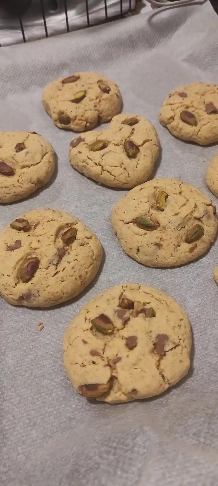 Several golden-brown cookies studded with green and brown pistachios sit on parchment paper, with one cookie in the…