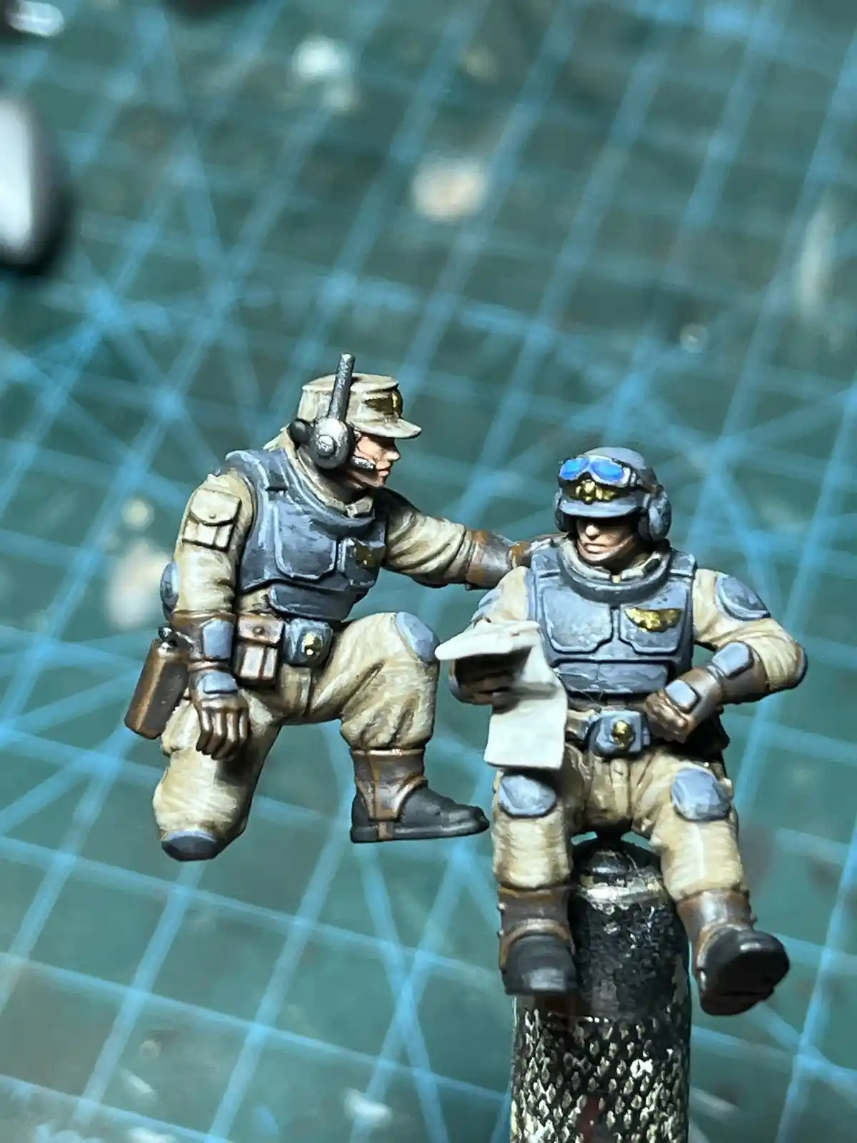 Two painted miniature soldiers in desert combat gear are posed on a textured surface; one kneels beside the other who sits…