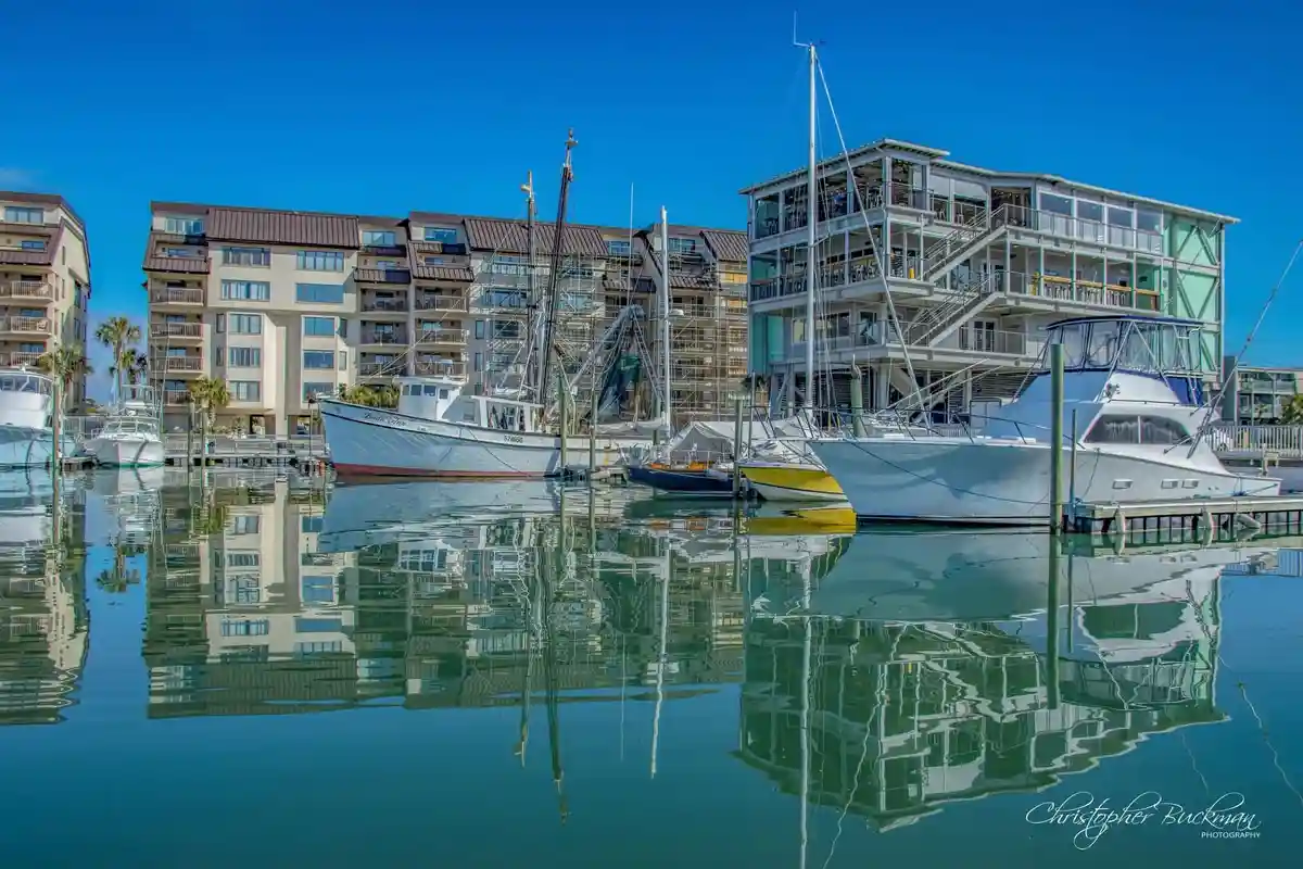 A serene marina scene with several boats docked in calm water, their reflections clearly visible. Multi-story buildings with…