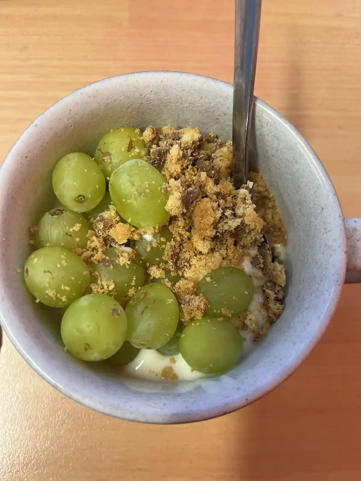 A top-down view of a speckled ceramic bowl filled with yogurt, green grapes, and granola, with a metal spoon inserted. The…