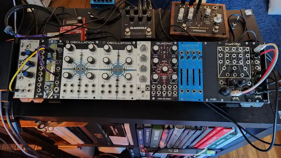 An overhead shot of a densely wired modular synthesizer setup on a black shelf, featuring multiple Eurorack modules like…