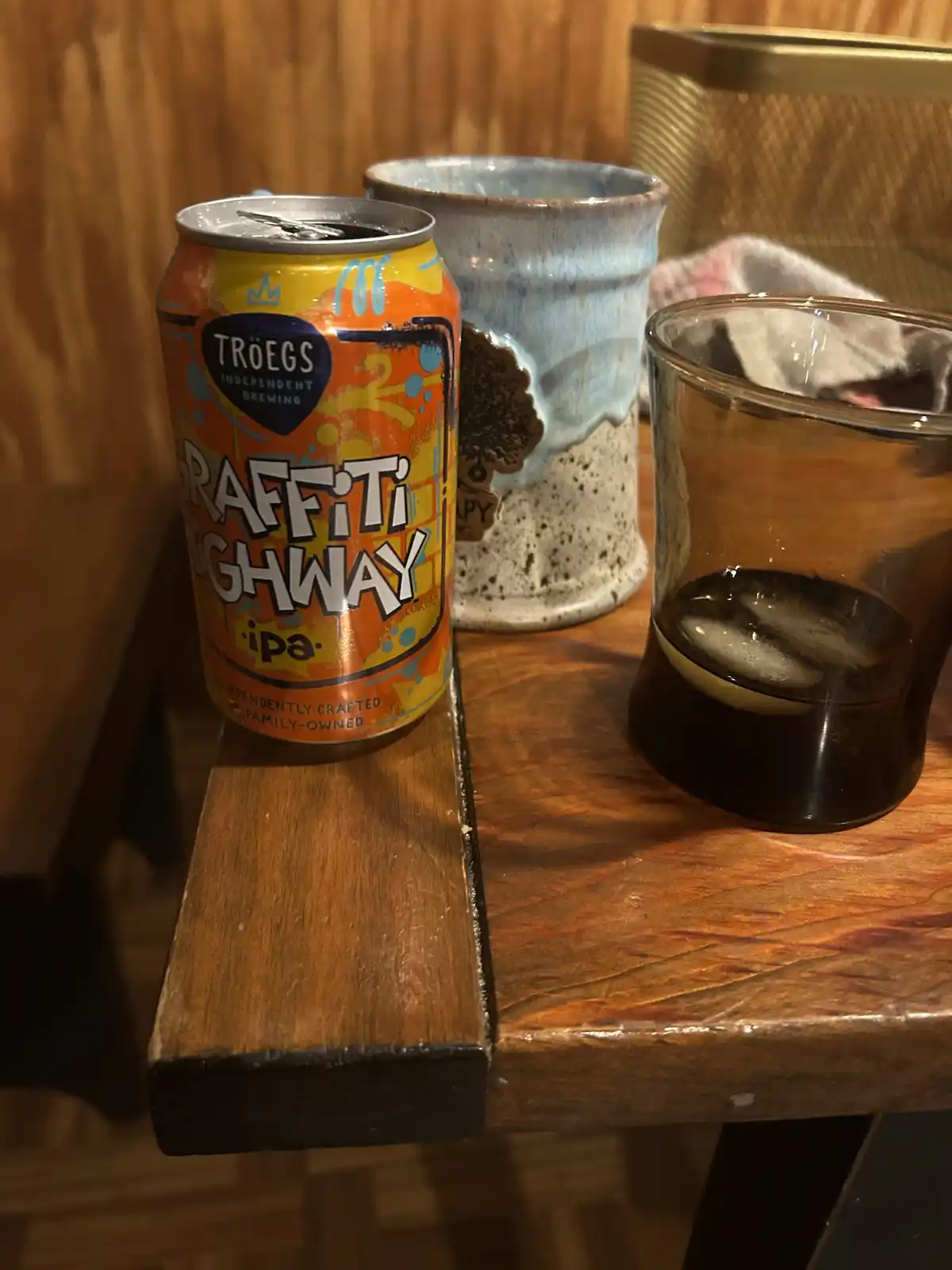 A can of Troegs Graffiti Highway IPA sits on a wooden table next to a glass of dark liquid with ice and a ceramic mug. The…