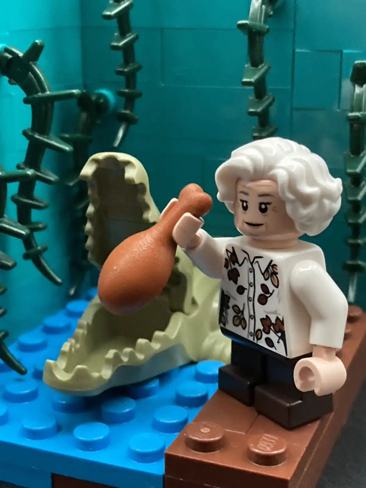 A lego minifigure with white curly hair and a floral shirt stands on a brown brick, holding a chicken leg. Behind her is a…