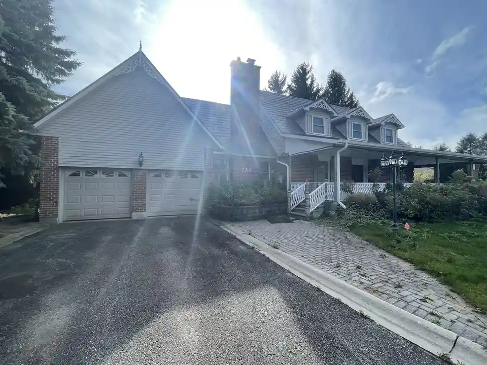A bright, sunlit photo of a two-story suburban house with white siding, brick accents, a double garage, and a front porch…
