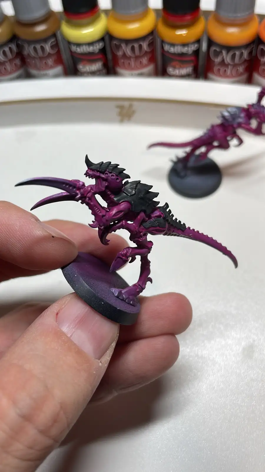 A hand holds a small, intricately painted purple and black fantasy creature miniature on a round base, with another similar…