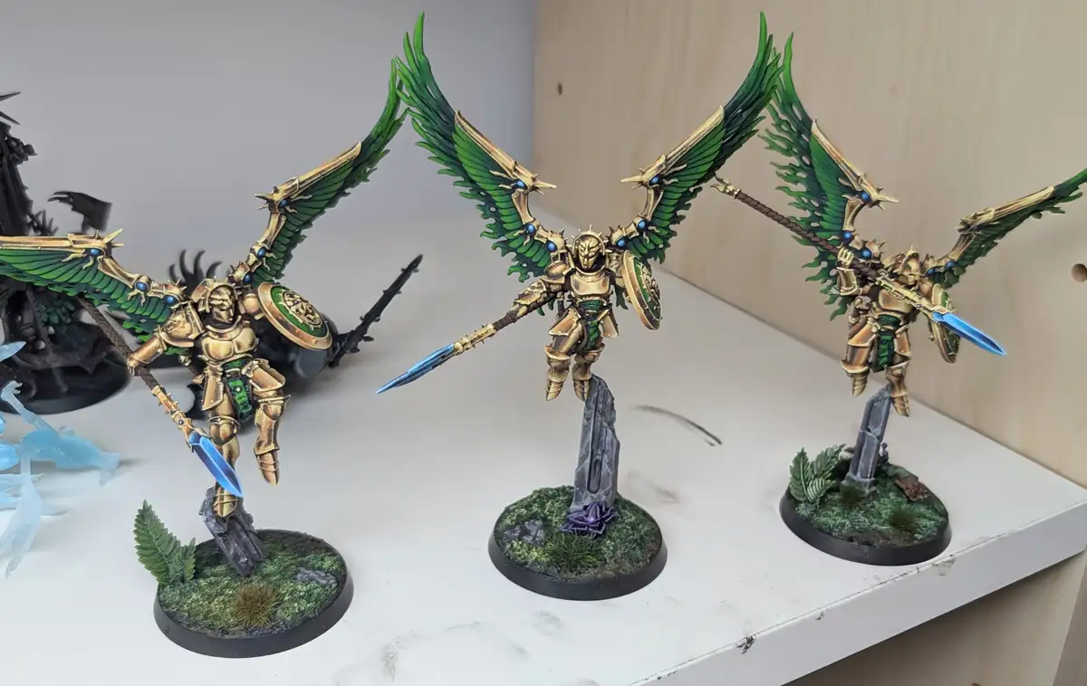 Three intricately painted miniature figurines, resembling armored angels with large green and gold wings, stand on detailed…
