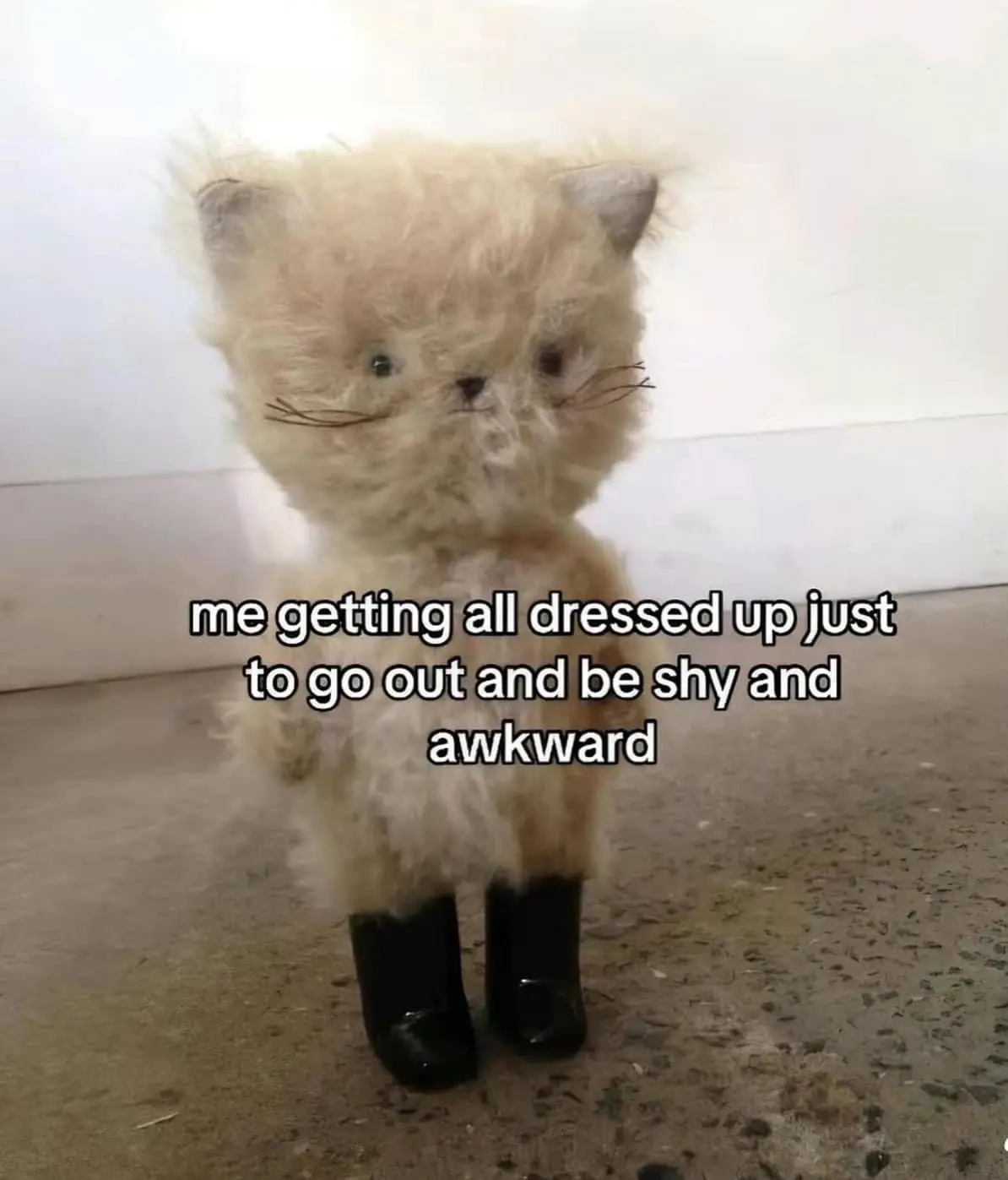 A fluffy, light-colored stuffed cat toy stands upright on a speckled floor against a plain wall, wearing small black boots…