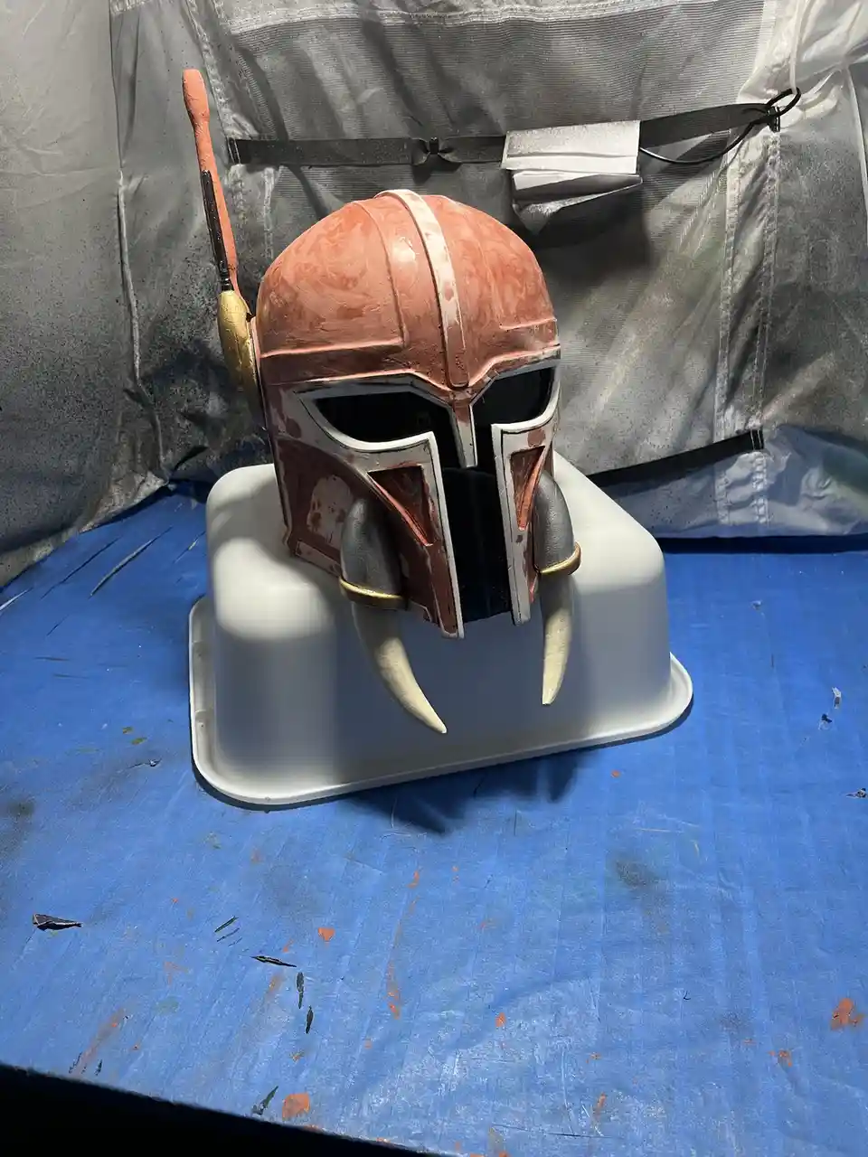 A partially painted Mandalorian helmet, resembling The Mandalorian from Star Wars, sits on a white plastic stand atop a blue…