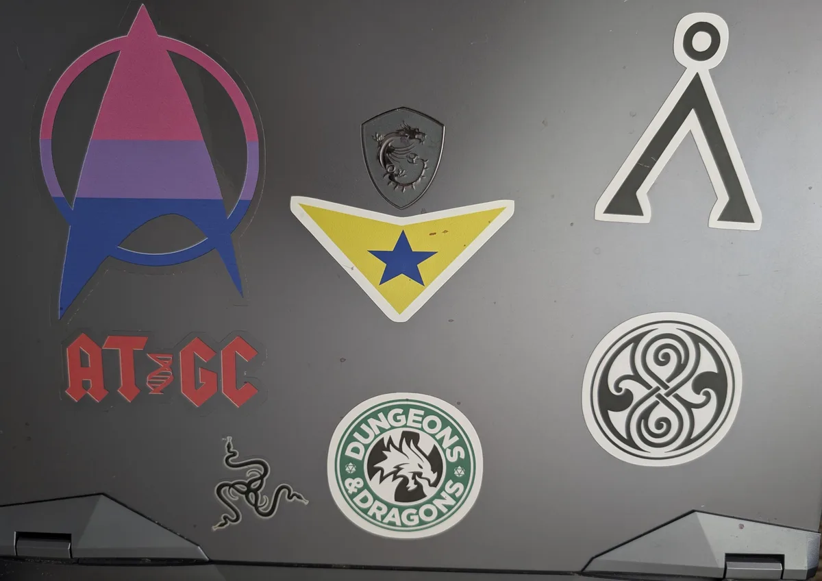 A close-up of a laptop lid covered in various stickers, including a rainbow-colored Avengers 'A' logo, a yellow triangle…