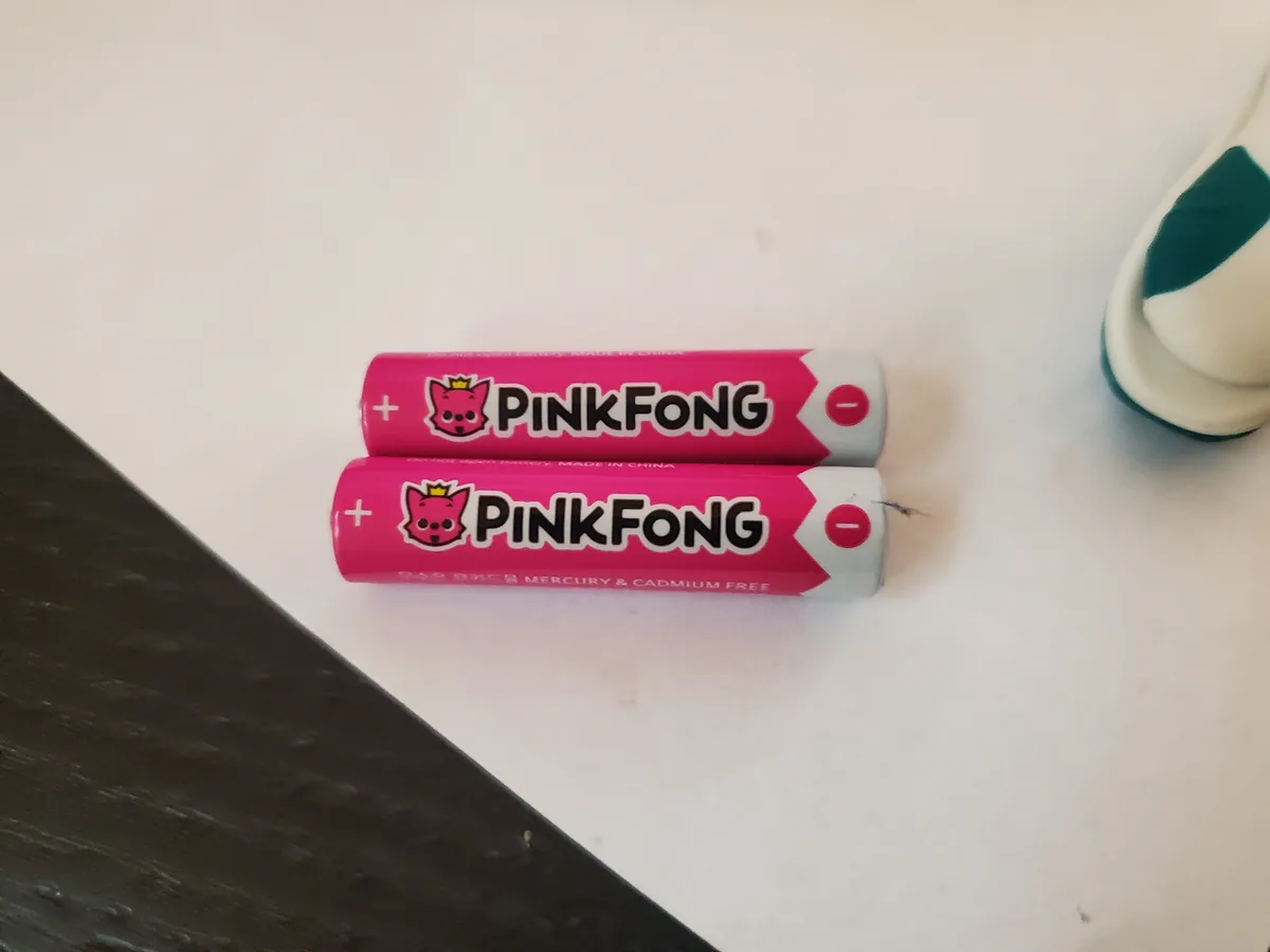 Two pink cylindrical batteries with 'PINKFONG' branding and a cartoon pig logo lie side by side on a white surface. The…