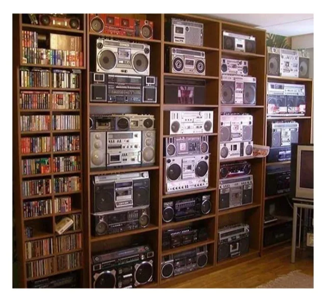 A room filled with wooden shelves displaying dozens of vintage boomboxes and cassette players, alongside bookshelves packed…