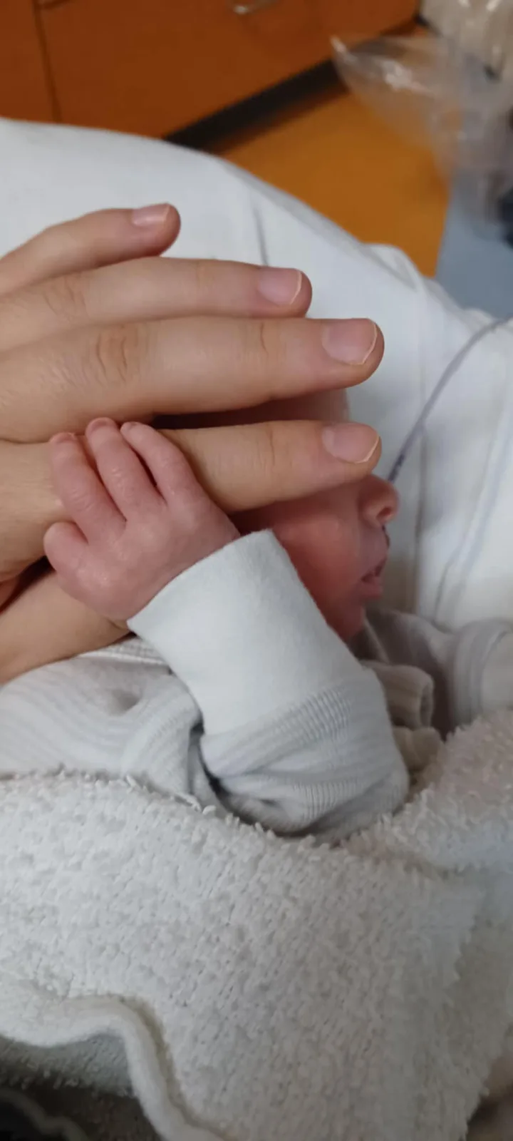 A close-up photo shows a newborn baby wrapped in a white blanket, with an adult's hand gently resting over the baby's face…