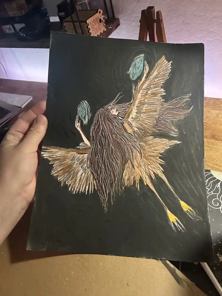 A hand holds up a dark-toned drawing of a winged angel with long hair, who is holding swirling orbs. The angel's wings are…