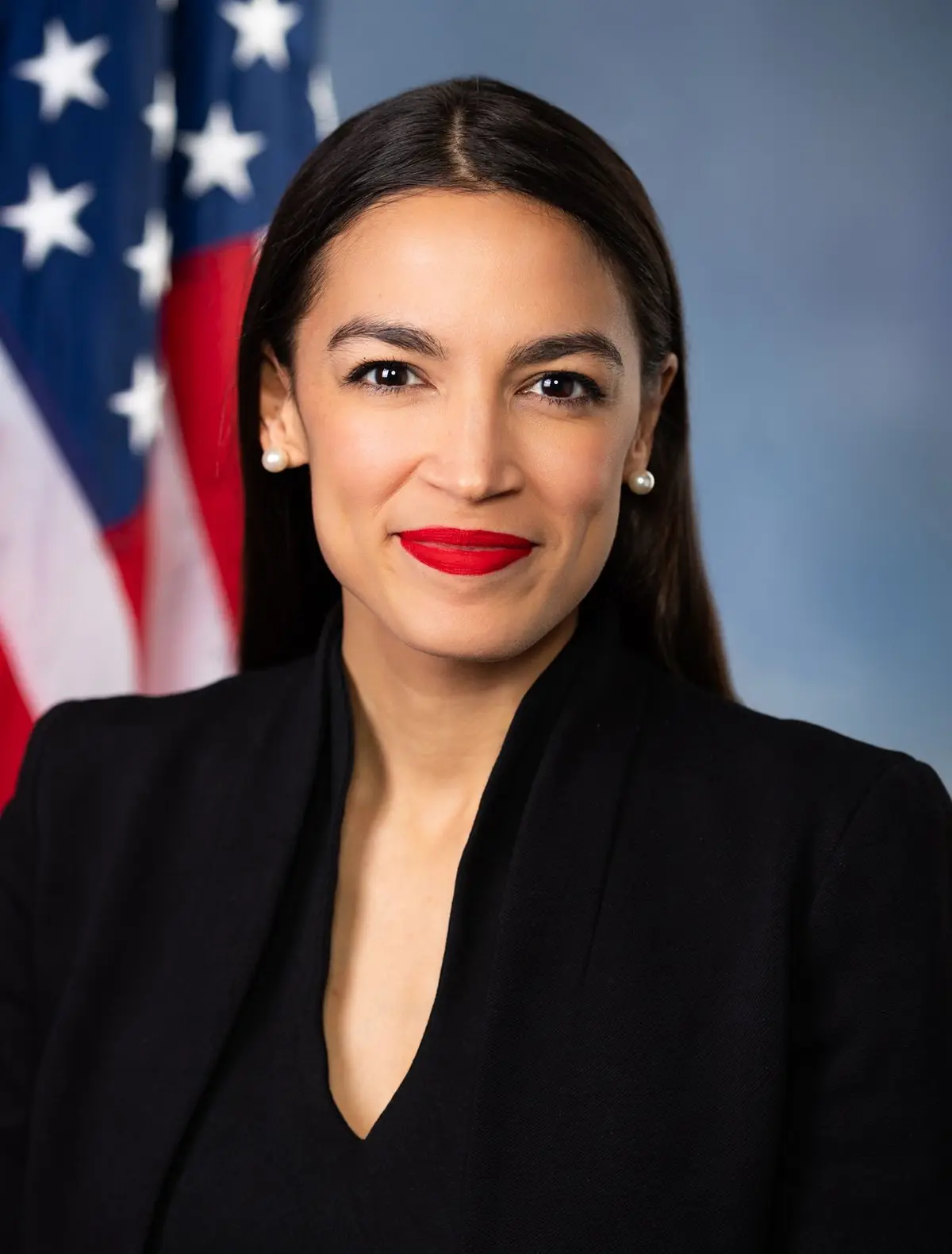 A formal portrait of a woman with dark hair, wearing a black blazer and pearl earrings, standing in front of an American…
