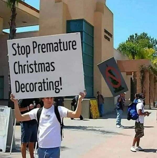 A person in a white t-shirt and backpack holds a large white sign that reads 'Stop Premature Christmas Decorating!' outdoors…