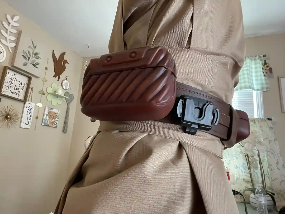 A person wearing a tan robe and a brown belt with a black, stylized lightsaber hilt buckle, with a brown quilted pouch…