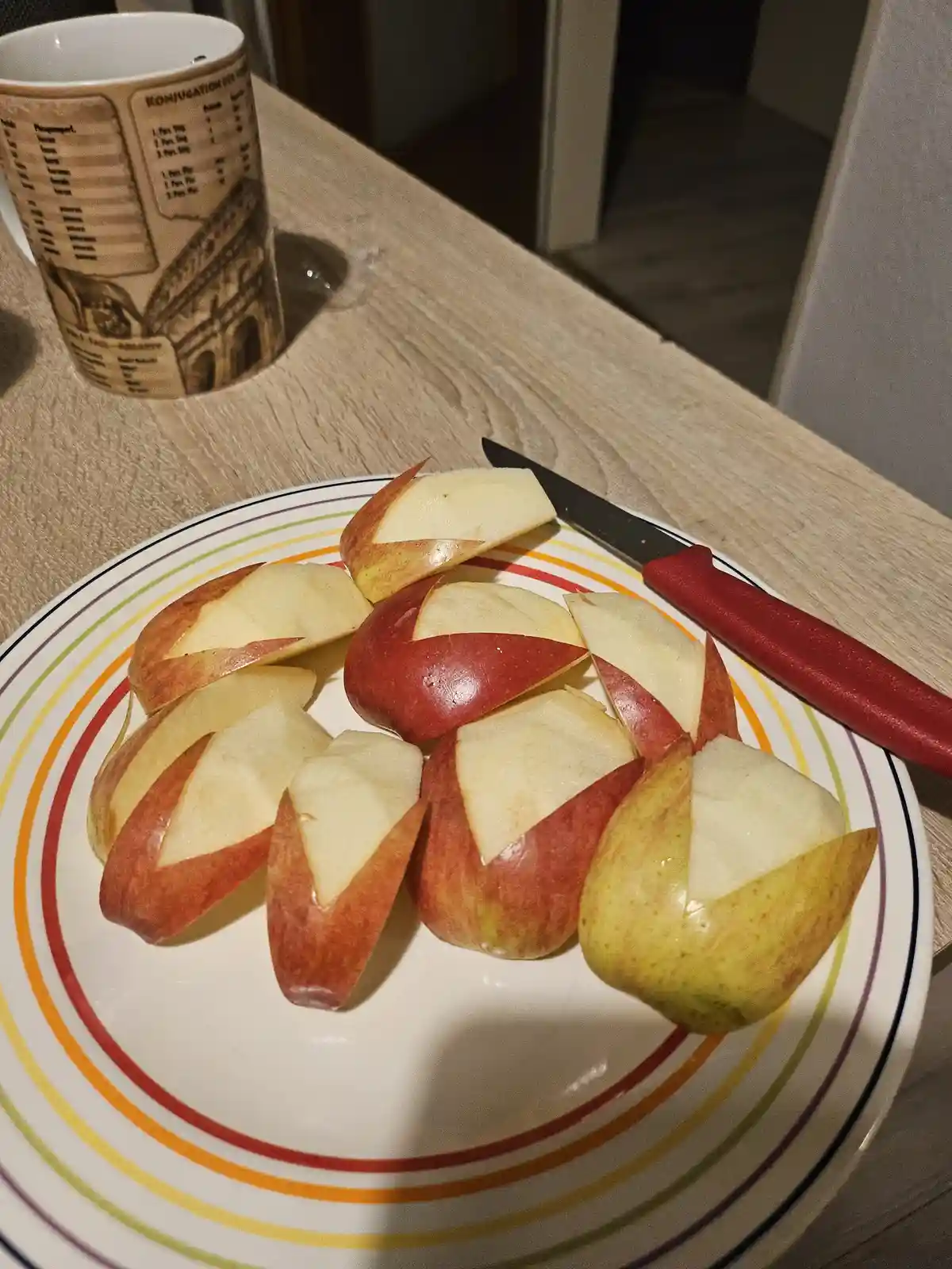 A plate holds several sliced apples arranged to look like a whole apple, with a knife and a mug featuring a 'Konjugation'…