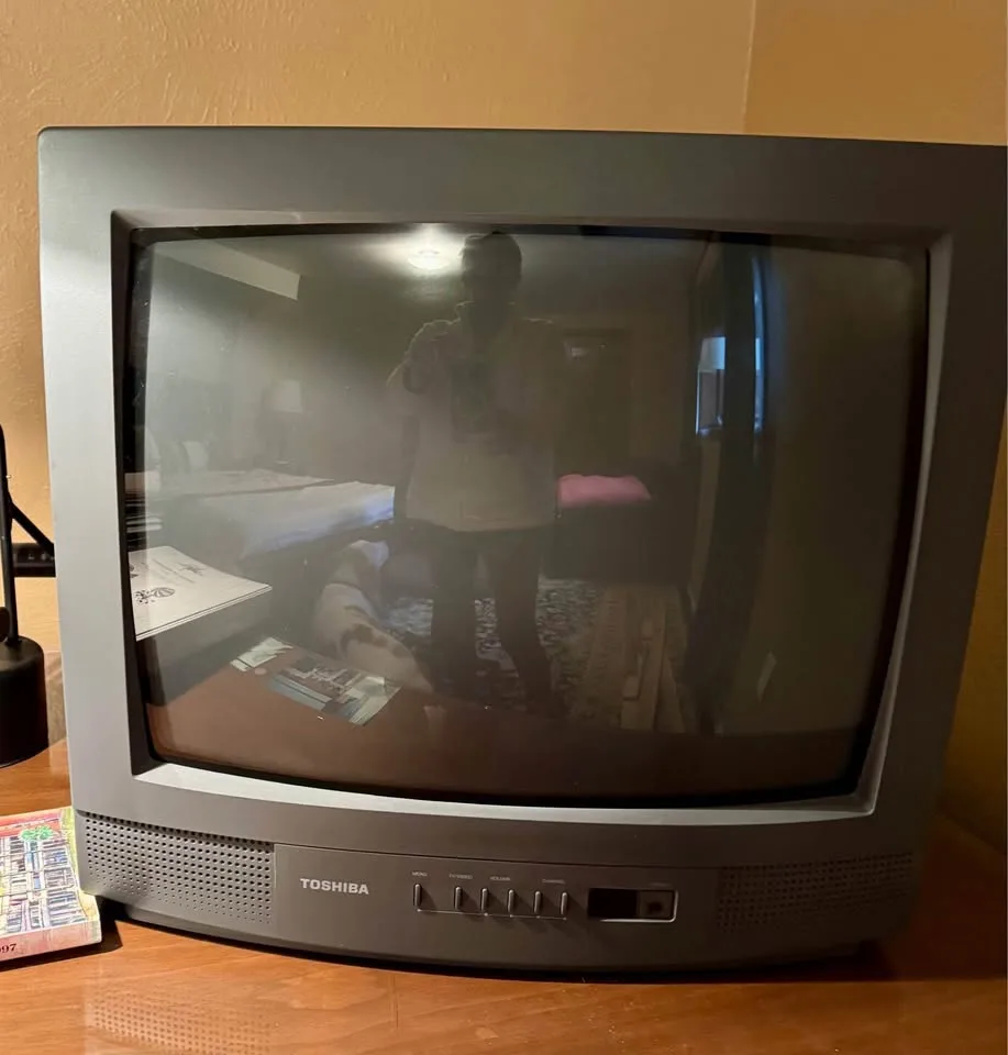 A person takes a selfie in the reflective screen of an old Toshiba CRT television, showing their reflection and a room with…