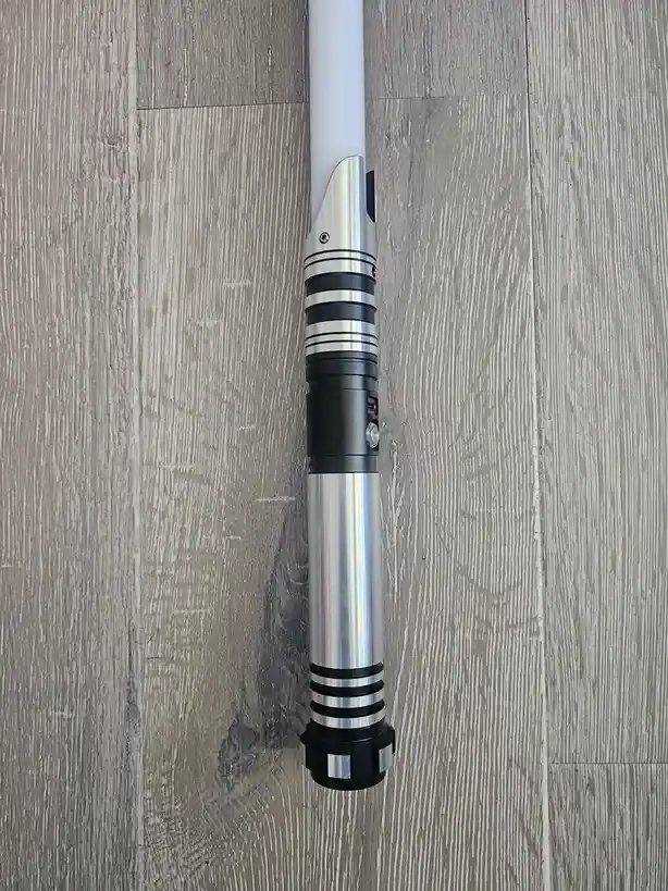 A white lightsaber hilt with silver and black accents lies vertically on a gray wood-grain floor. The overlaid text reads…