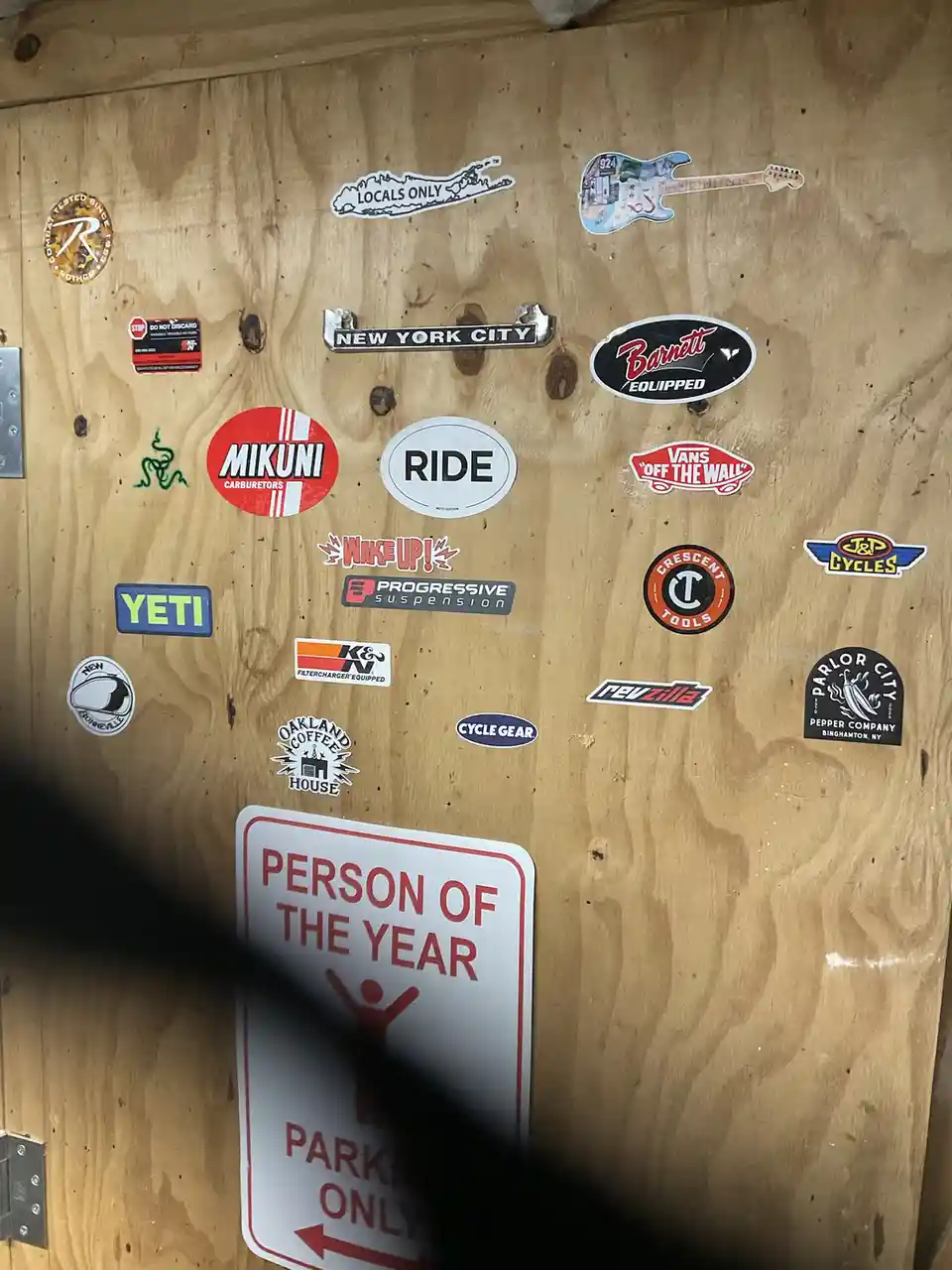 A wooden surface covered in various stickers including 'New York City', 'Vans', 'YETI', 'Mikuni', and 'Person of the Year'…