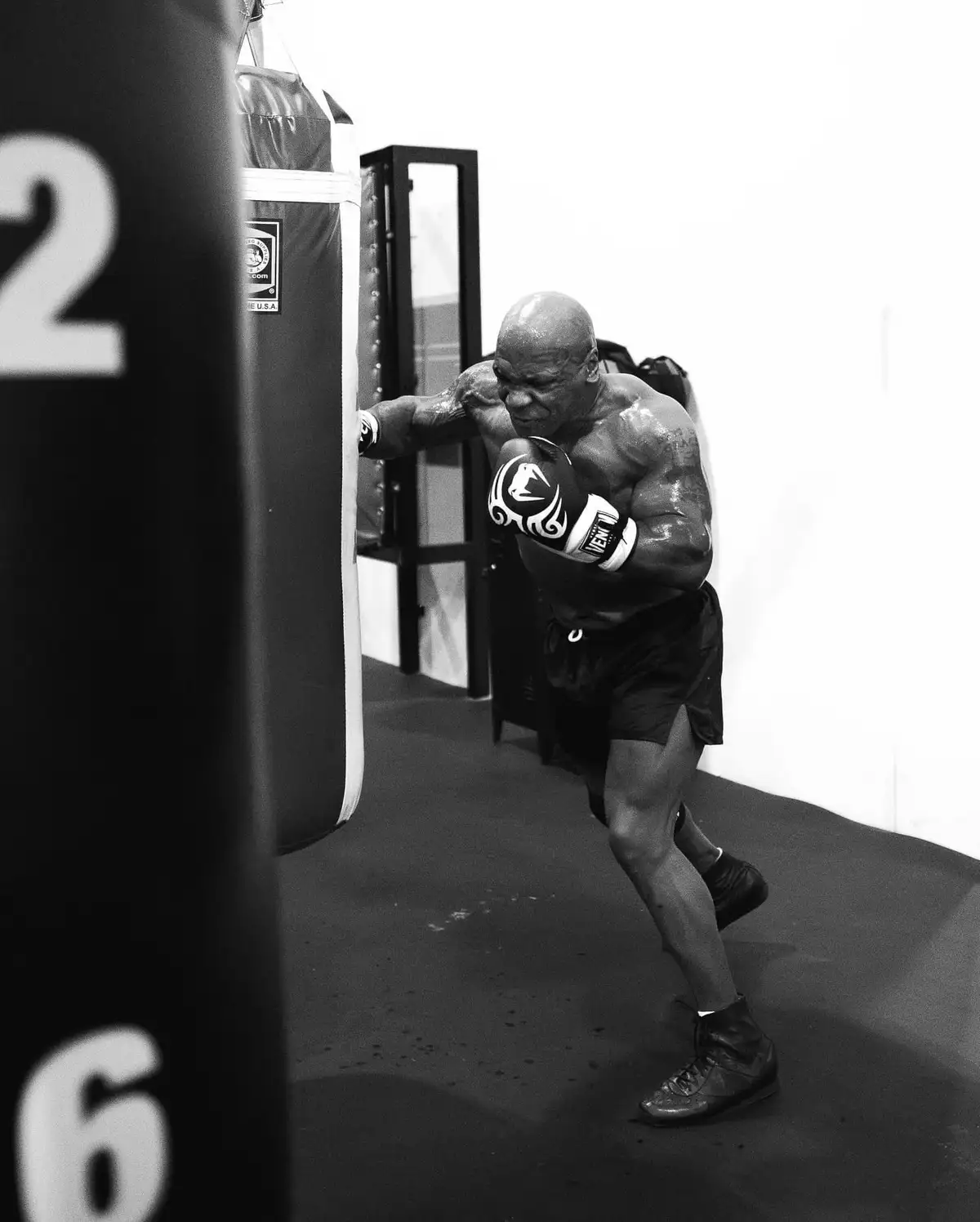 A black and white photo shows a muscular, bald man in boxing gloves and shorts, mid-punch on a heavy bag in a gym. The image…