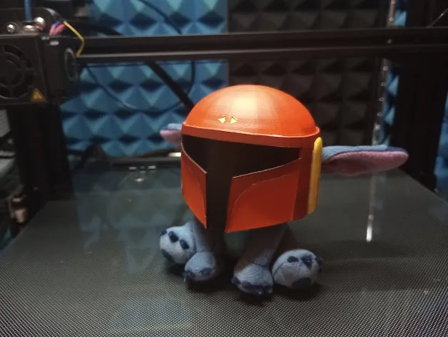 A plush Stitch toy is sitting upright on a 3D printer bed, wearing a red Mandalorian helmet with yellow markings. The…